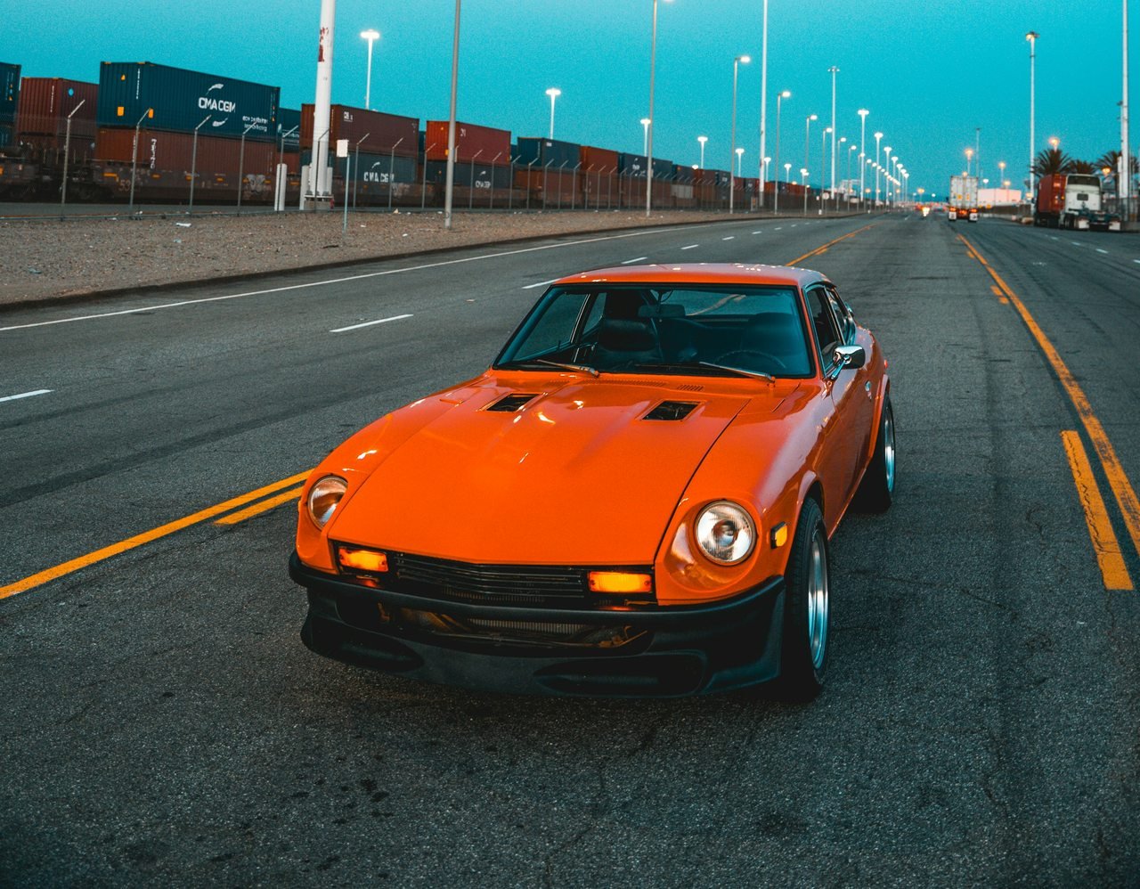 Orange vintage Japanese sports coupe on a broad road at dusk.